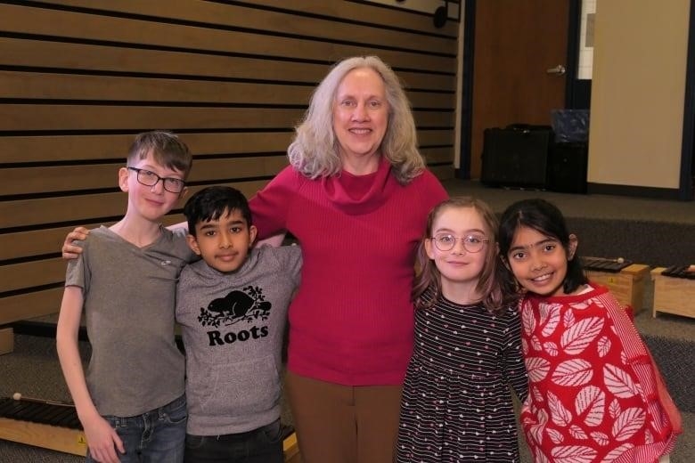 A teacher stands to the right of students, everyone playing ukeleles and singing, in a music classroom. A large treble clef and musical notes decorate the wall behind her.