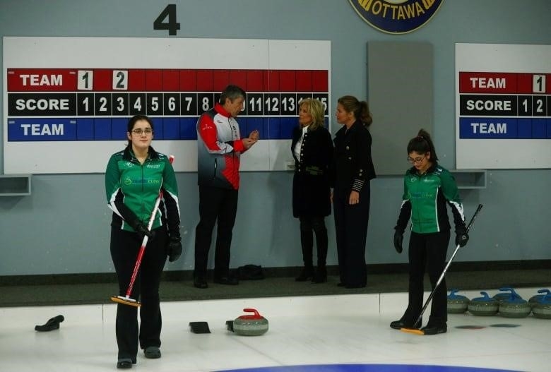 Two women listen to someone talk about curling next to a rink.