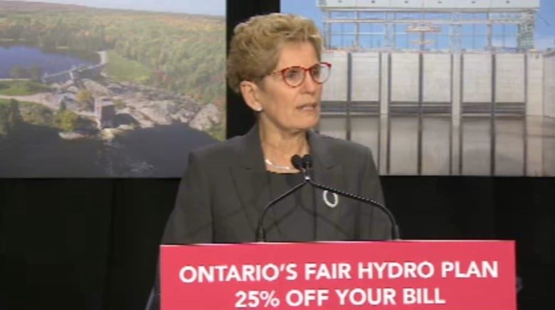 Kathleen Wynne stands at a podium that reads "Ontario's Fair Hydro Plan 25% Off Your Bill." 