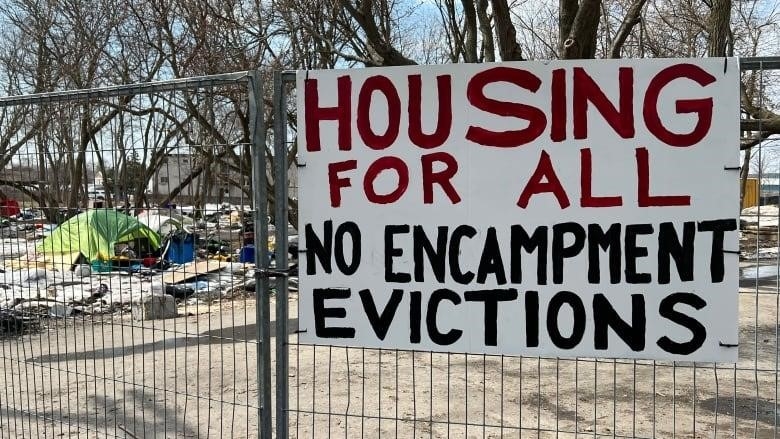 A large white board is painted with the message, "Housing for all, no encampment evictions." It's hanging from a fence and a tent can be seen on the snowy ground behind it.