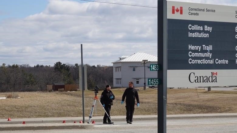 A police officer in a bright yellow jacket ties yellow caution tape to a light post. A cruiser and a sign for the Collins Bay Institution can be seen in the background.