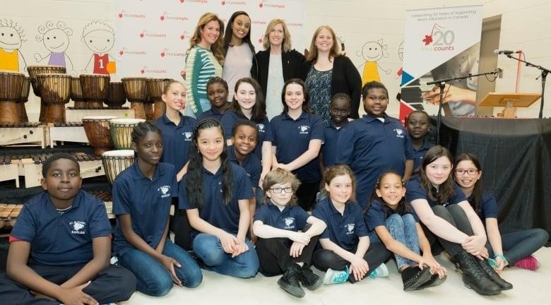 Four smiling adult women stand behind a group of 16 sitting and kneeling elementary students, alongside bright signage, drums and other percussion instruments.