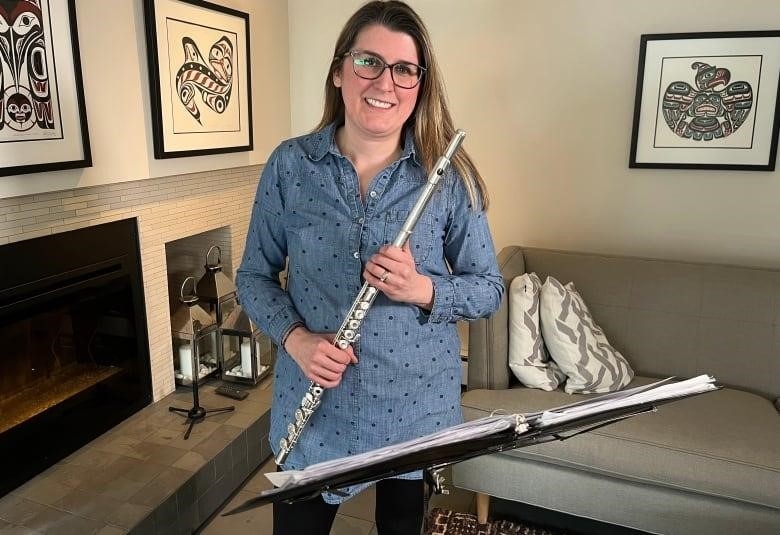 A smiling woman in glasses, a denim shirt and leggings holds her flute while standing behind a music stand, with a family living room seen behind her.