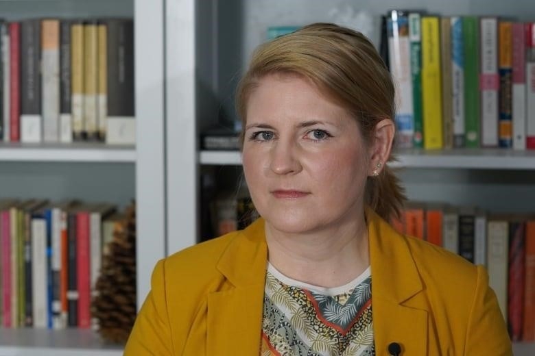 A woman in a mustard-coloured blazer and patterned shirt, with her hair pulled back,  stands before a wall of full bookcases. 