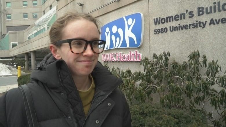 A blond woman wearing tortoiseshell glasses and a black puffy coat stands outside the IWK Health building in Halifax, Nova Scotia.