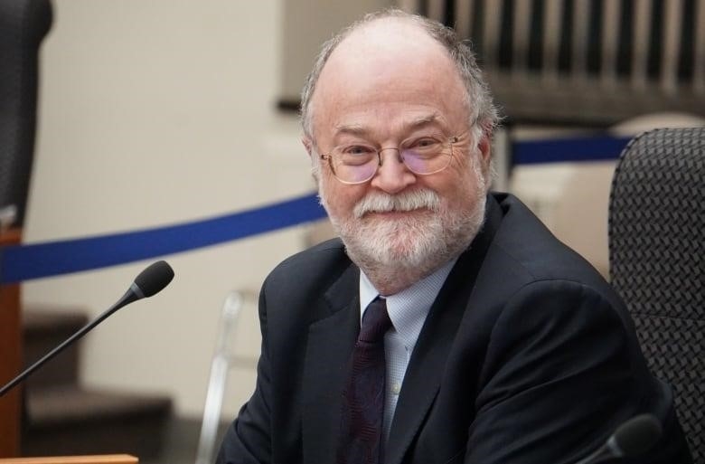 A seated man with white-grey hair and facial hair, wearing a suit, smiles at the camera from behind a microphone.