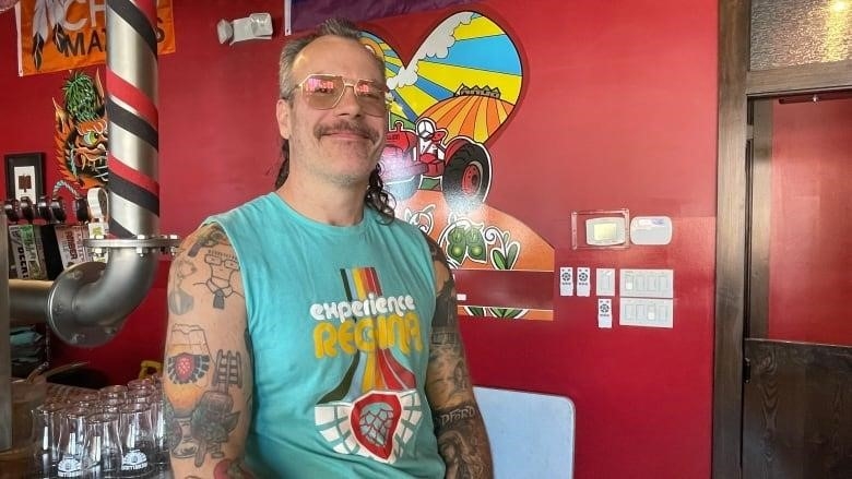 A man in a mullet and glasses leans against a counter. The man is wearing a turquoise blue armless shirt with the phrase "Experience Regina" on it. 