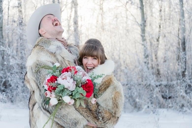 A smiling couple stands outside on a cold winter day wrapped in fur jackets. 