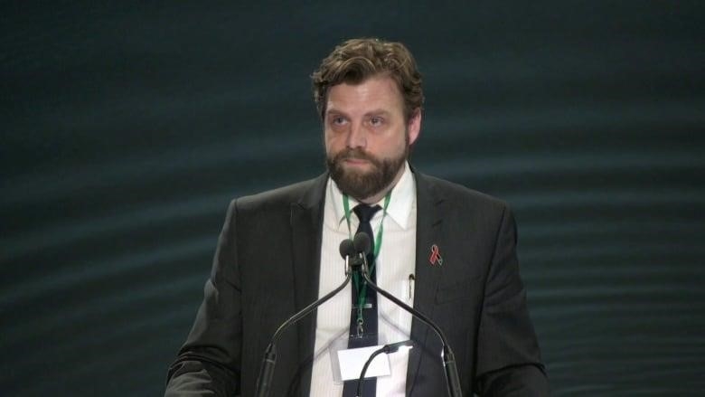 A white man with brown hair and beard wears a suit with black jacket and white shirt, while standing at a podium with a microphone.