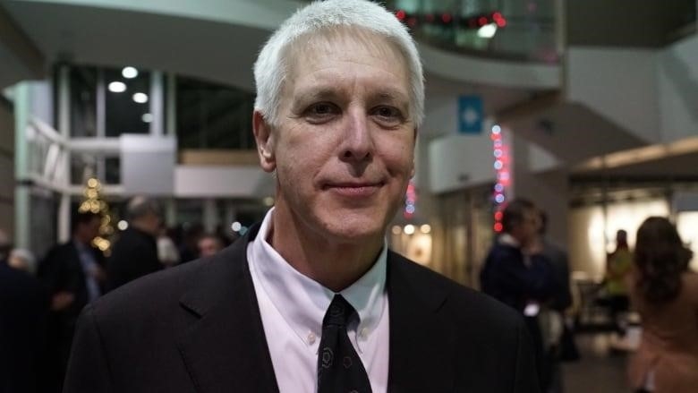 An older man with white hair and wearing a suit looks at the camera.