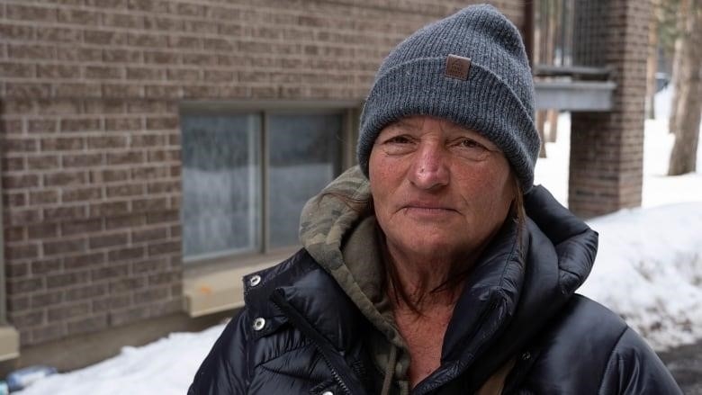 A woman with a toque and winter jacket stands on a path bordered by snow near an apartment coplex.