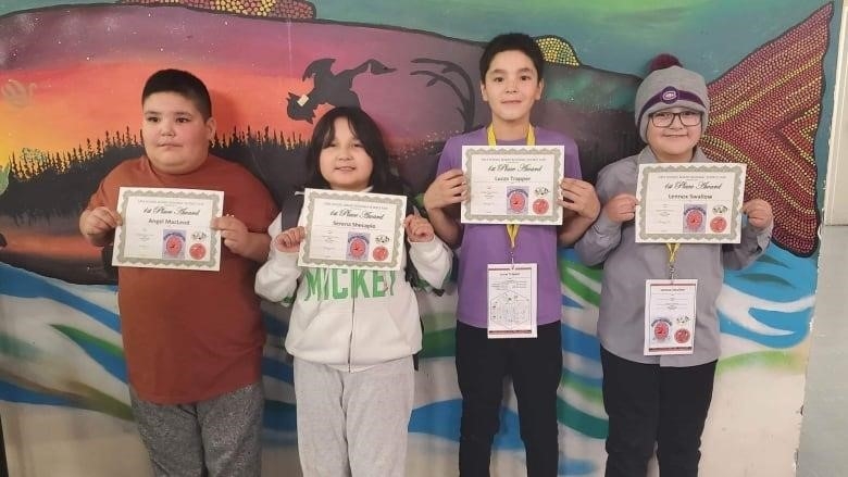 a group of Indigenous grade five students stand against a colourful wall with a fish painted on it. They hold certificates their science project has won.