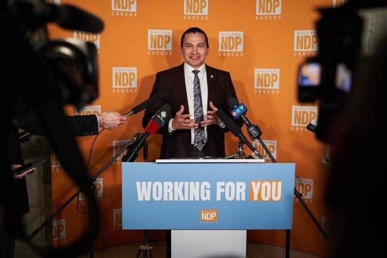 A smiling man in a suit stands at a podium behind a number of microphones, with a backdrop behind him with the Manitoba NDP logo.