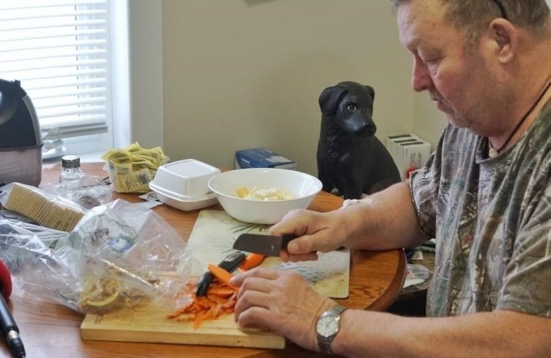 A man holds a knife and is cutting carrots. There's a pile of carrot and turnip peels on the table wooden cutting board. 