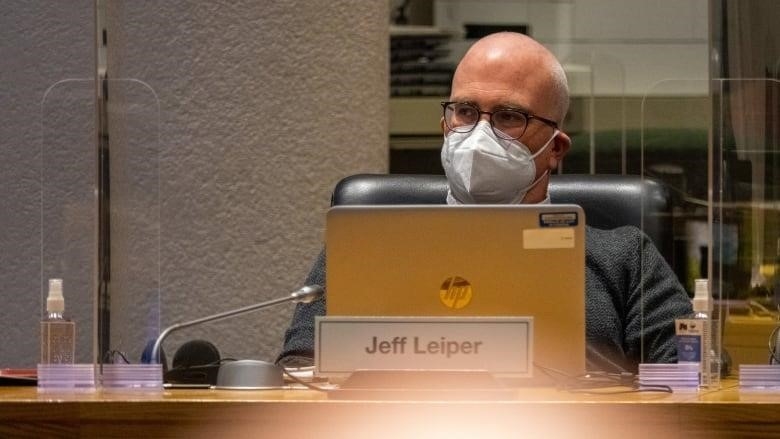 A man in a mask sits behind a laptop at a table.