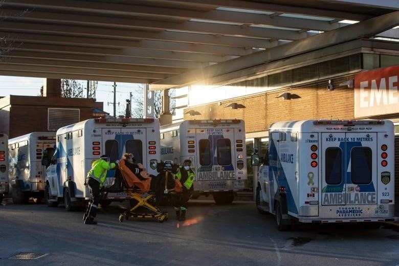 Paramedics push a patient on a stretcher towards the door of an emergency department as four other ambulances are shown parked by the entrance.