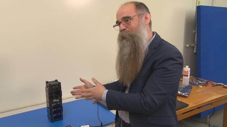 A man with glasses and a long grey and brown beard gestures to a small black tower on a table.