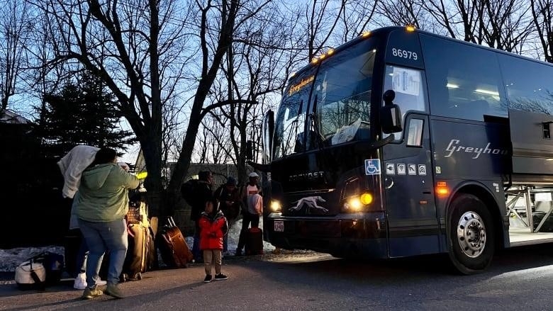 A woman carries a baby. A toddler looks at the bus. A group of people wait in line to get on a Greyhound bus as the sun sets. 