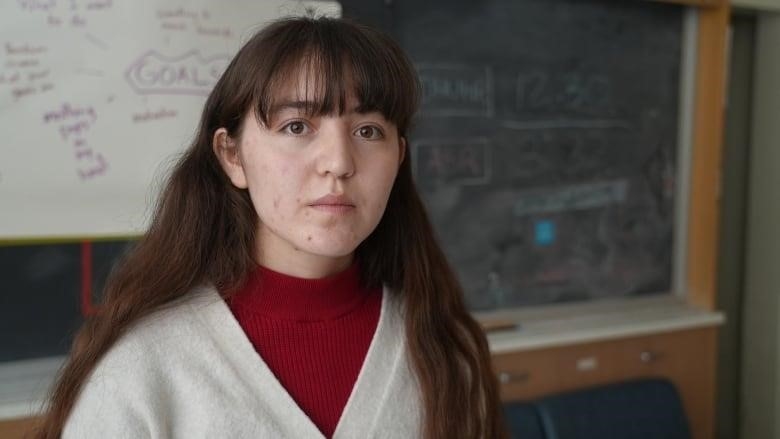 A solemn teen wearing a cream sweater and red turtleneck stands in a school classroom, with a blackboard and whiteboard scrawled with writing seen behind her.