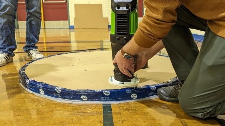 A circular unit with plastic film taped to the bottom and a leaf blower strapped to it sits at one edge of a gymnasium with a crowd looking onward at it