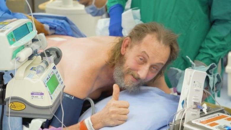 A man lying on his stomach on an operating table gives the thumbs up for the camera.