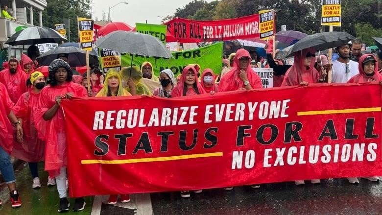 Many people wearing raincoats and holding umbrellas stand behind a red banner.
