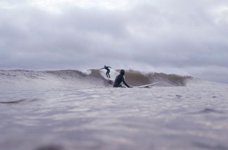 A surfer rides a wave in the distance, while a person wearing a wetsuits sits on a surfboard in the foreground.