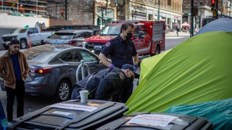 Two firefighters wearing masks peer into a tent on a city sidewalk, with an onlooker and queued-up cars behind them.