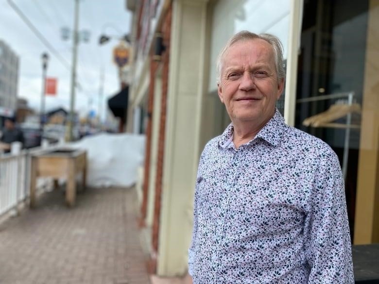 A man in a collared shirt looks stands in front of a building. Behind him is a small fenced in area used for outdoor seating in the warmer seasons. 