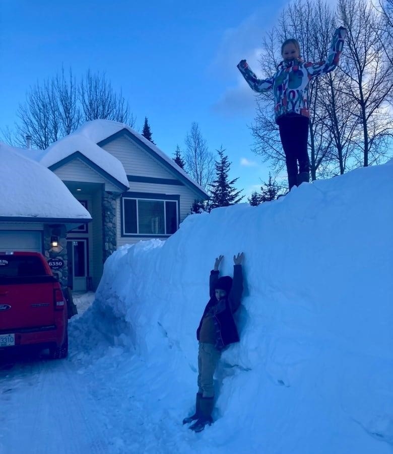 One girl stands on top of a snow pile while another leans into it.