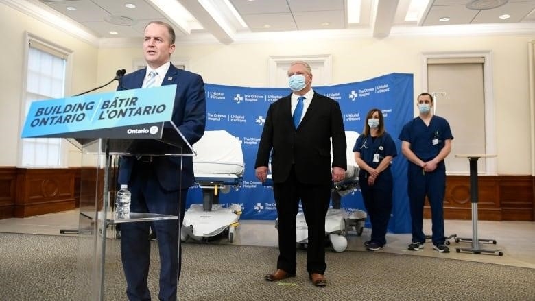 A man in a suit speaks at a podium while another man in a suit looks on.