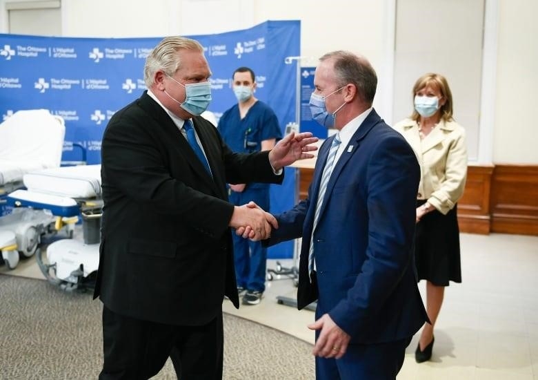 Twp men wearing suits and masks shake hands at a hospital news conference.