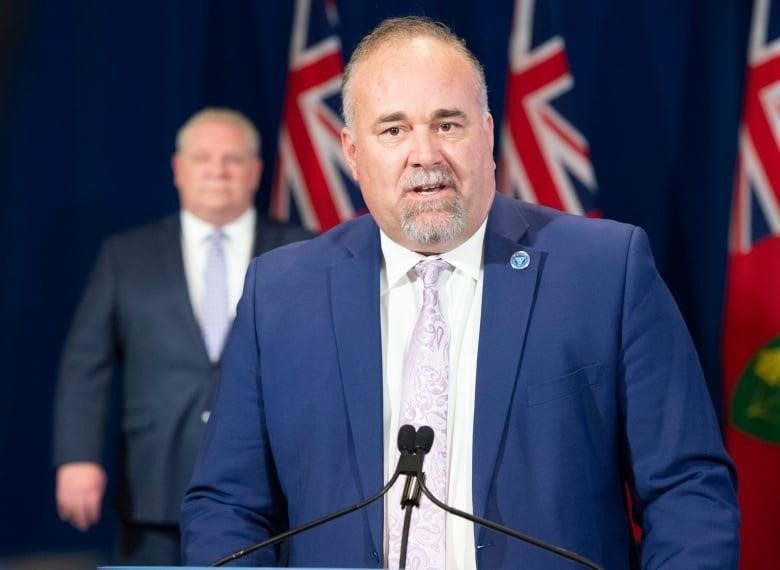 Energy Minister Todd Smith stands at a podium during a news conference while Premier Doug Ford looks on from behind him.