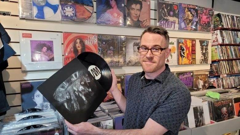 A man is shown in a record store holding a piece of vinyl being pulled out of the cover with a number of albums displayed behind him.