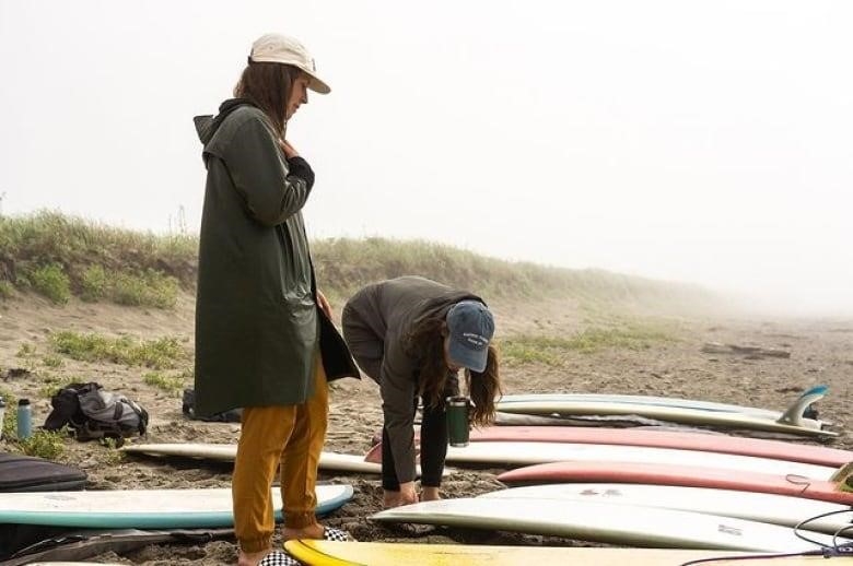 Two women stand next to a row of longboards on a sandy beach.