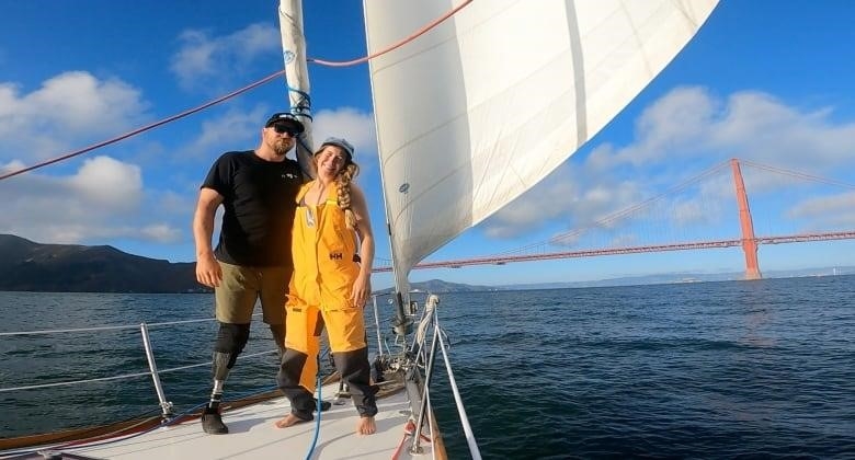 A man and a woman smile for a picture aboard a sailboat, with an orange bridge visible behind them.