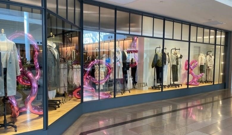 A pink, curved sculpture is pictured inside a window display for a store inside a mall. Women's clothing is pictured on nearby mannequins.