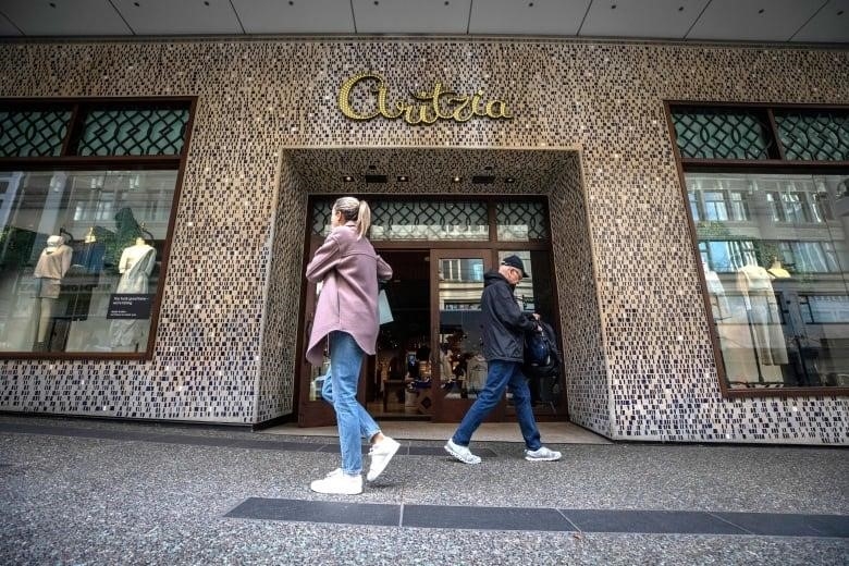 Two people wearing spring clothing walk in opposite directions past a storefront in downtown Vancouver. The word "Aritzia" appears over the wooden doors in gold metal script.
