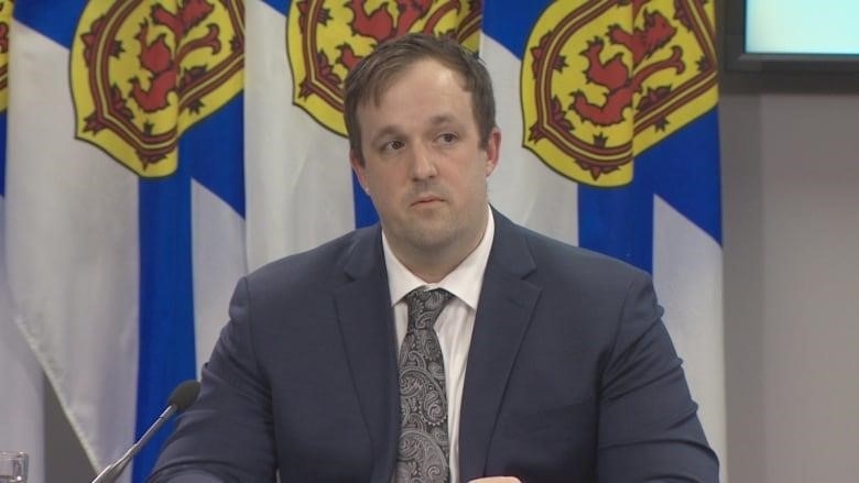 A white man in a suit sits at a table in front of a row of Nova Scotia flags