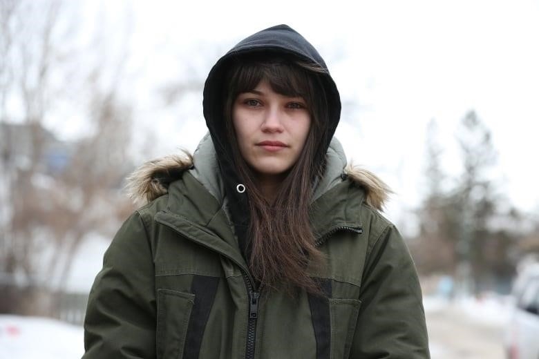 A woman with long brown hair wearing a black hoodie and an army green parka looks at the camera.