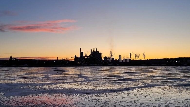 A mill with smokestacks silhouetted against a vibrant sunset is seen in the distance across a frigid body of water.