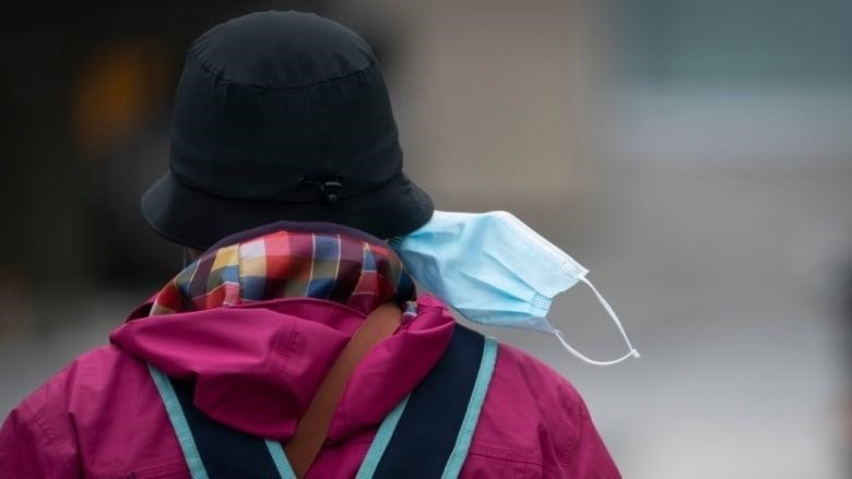 Back view of a woman wearing a hot pink rain coat with a multi-colour hood, a black hat and a backpack, walking with her medical mask half-on and blowing in the wind.