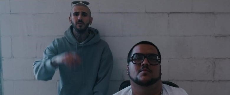 Two men are seen looking at the camera in front of a white concrete wall.