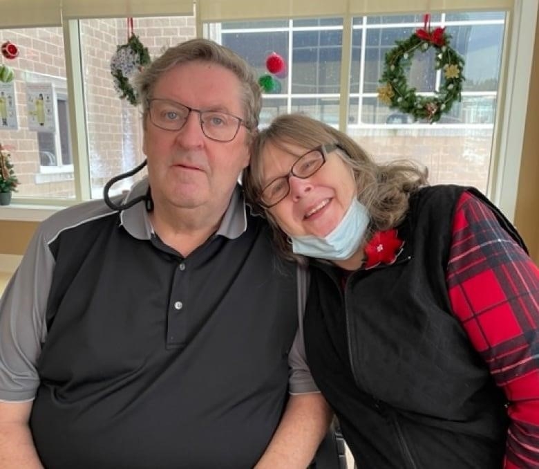 An older man and woman stand next to each other on the front porch of a house. The man doesn't wear a shirt, but suspenders, which he's stretching away from his chest. He has a mischievous smile on his face. The woman next to him is laughing.