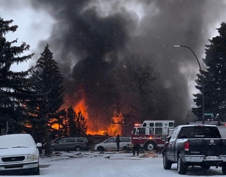 A fire engine and several vehicles surround an area where large flames are burning and sending thick black smoke into the snow-filled air.