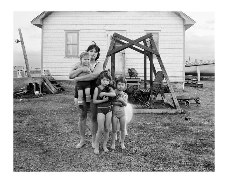 A black and white photograph of a woman standing in front of a house holding a young child, while two other children stand close to her. There is a large wooden swinging bench on a platform between them and the house.