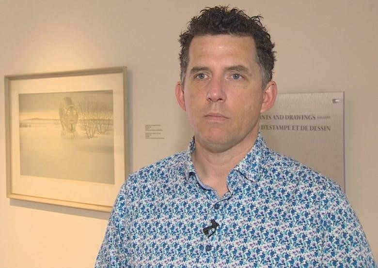 A man with short dark hair looks seriously at the camera. He is wearing a collared dress shirt with a blue and white pattern, and standing in front of a white gallery wall with art works hung on it.