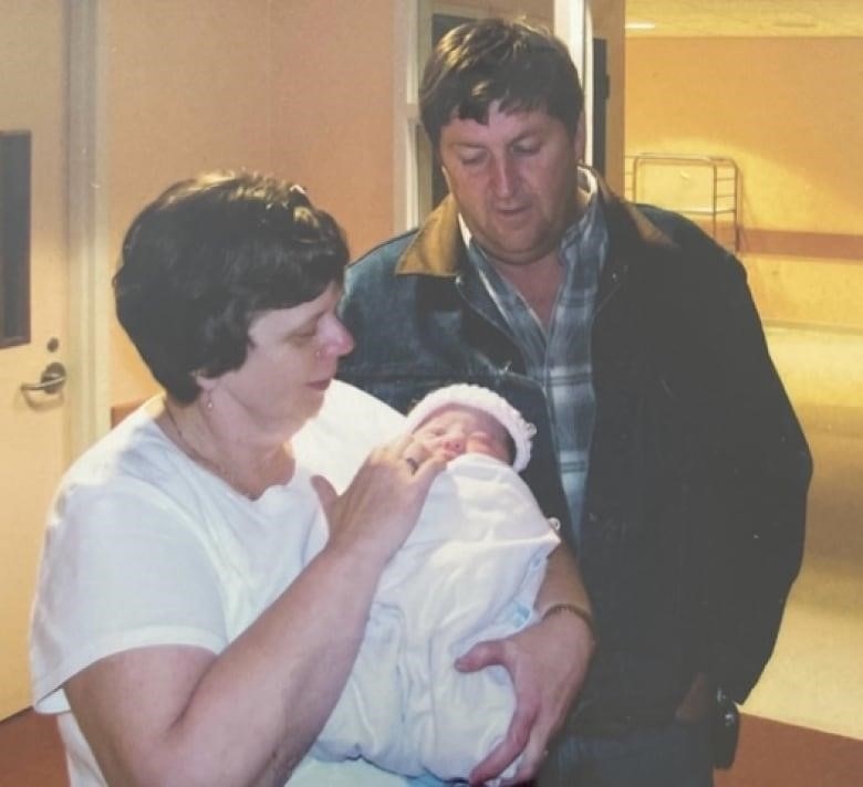 A woman is holding a baby, looking at it. A man is standing behind her, looking at the baby, as well.