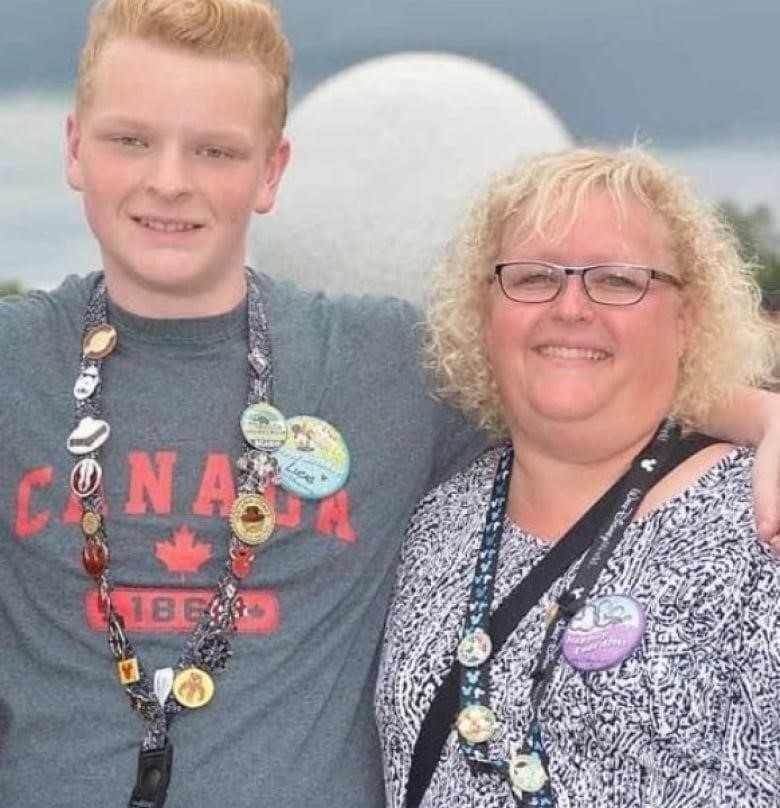 A white woman with glasses and black and white pattenered shirt poses with a young man with red hair and a grey teeshirt. The boy has his arm around the woman, and both are wearing lanyards with buttons.