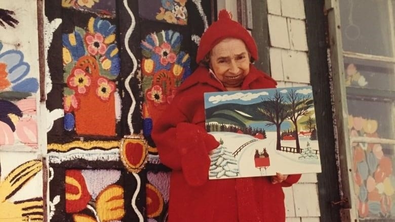 A petite artist is shown with a piece of her art work in front of a colourful house.